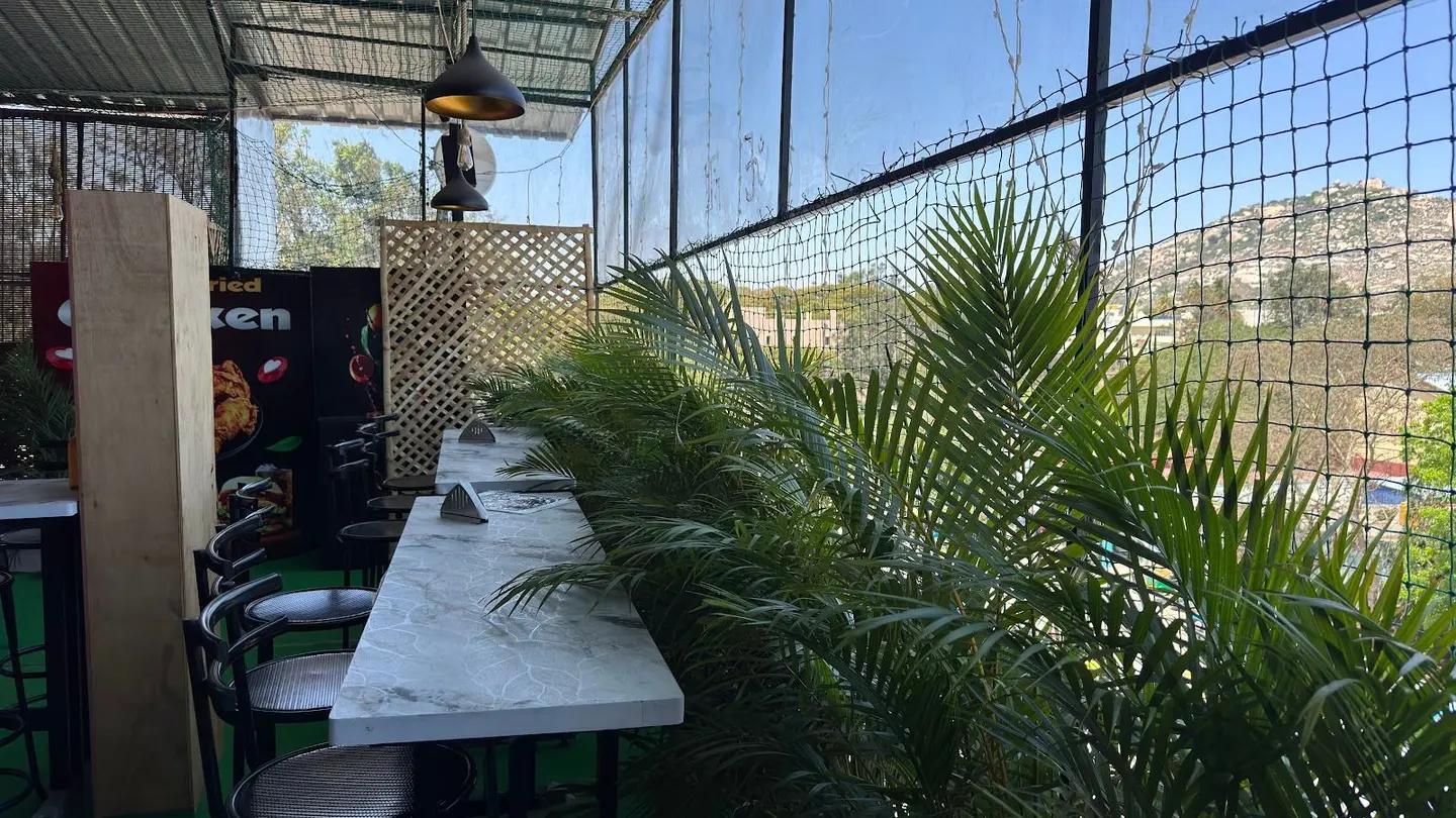 A corner of the roof: marble tables against a lattice screen, a black pendant lamp, potted palms, and the hill through the mesh.