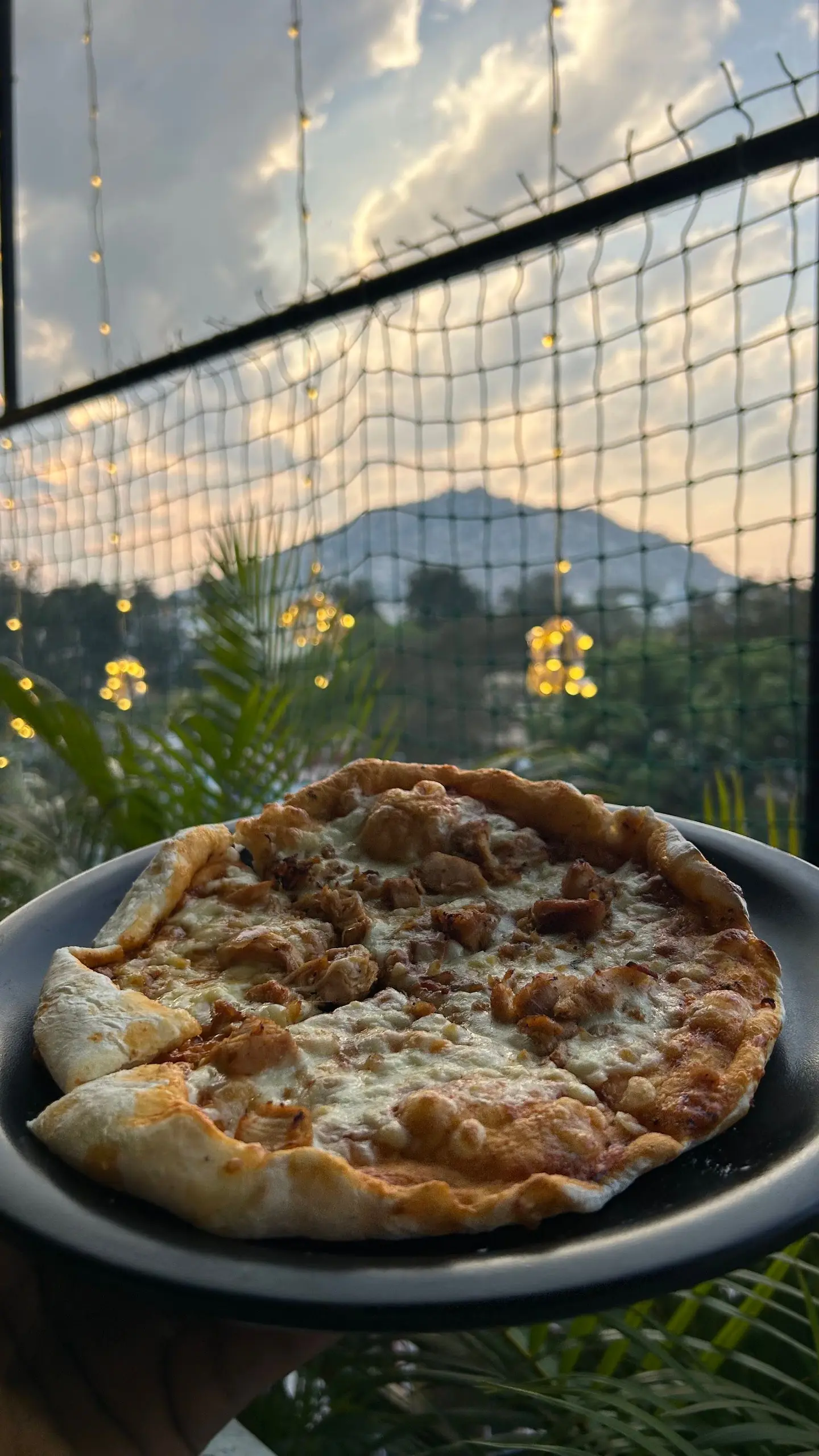 A whole pizza on a dark plate in the foreground with the hill and a pink dusk sky behind it.