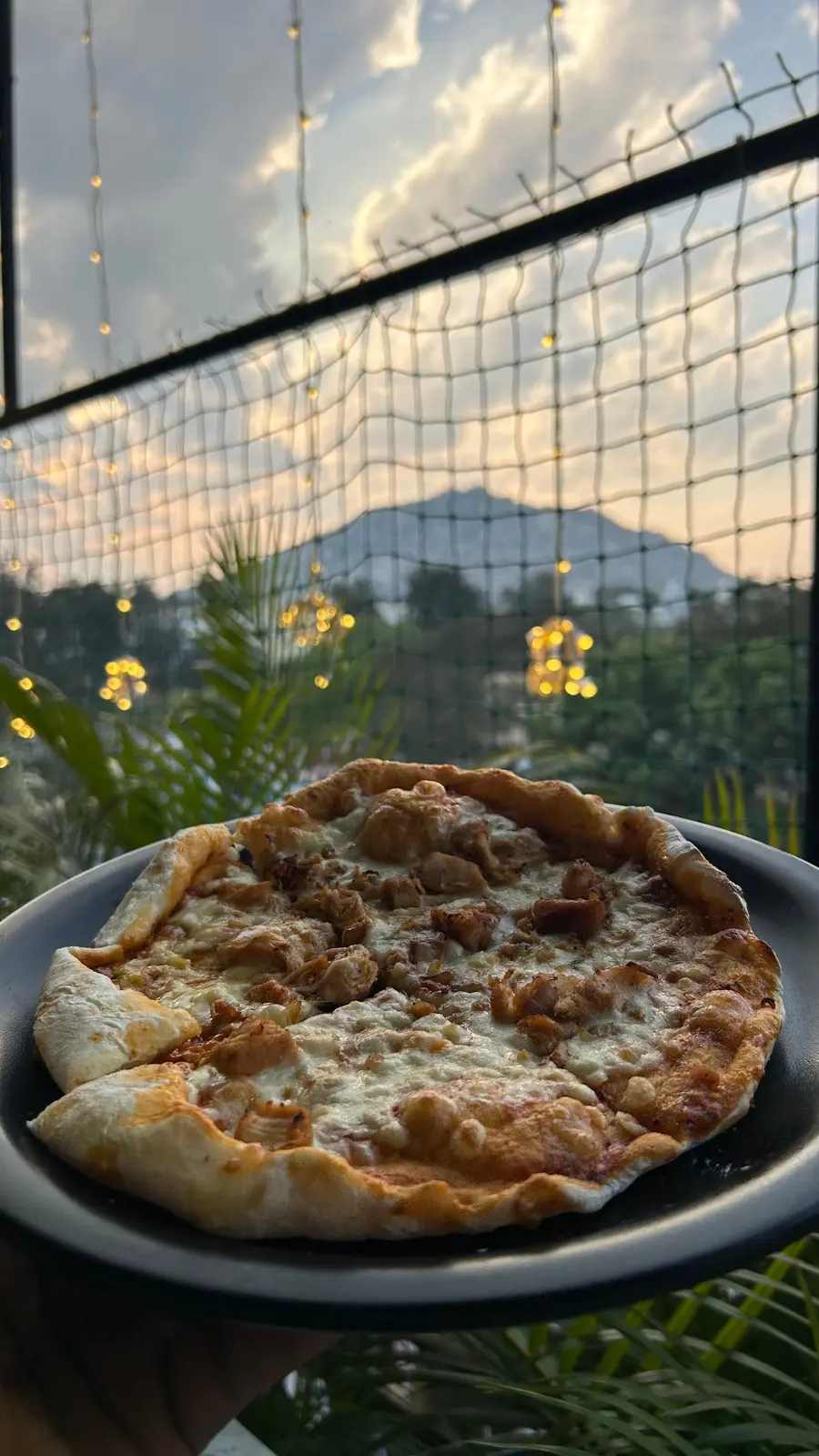 A pizza on a dark plate on the rooftop at dusk, with a hill on the skyline behind the wire mesh and fairy lights strung overhead.
