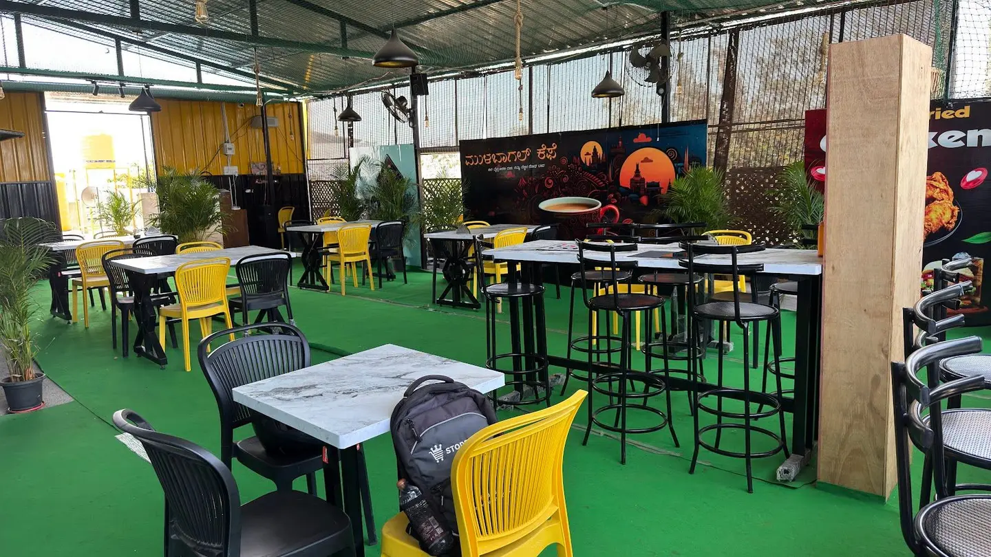 The rooftop under a steel shed roof: green turf floor, yellow chairs, marble-topped tables, tall stools and black pendant lamps, with the painted Kannada mural along the far wall.