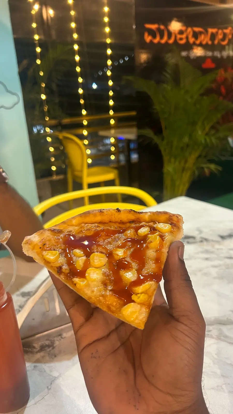 A hand holding a slice of sweetcorn pizza with a drizzle of sauce, fairy lights and a yellow chair behind.