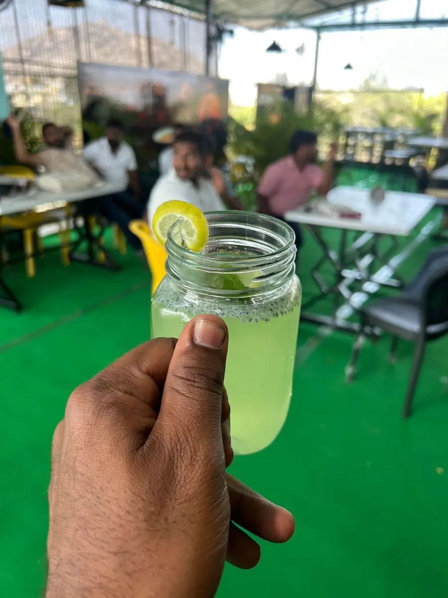 A hand holding a mason jar of cloudy lime soda with a slice of lemon in it, green turf and white tables behind.